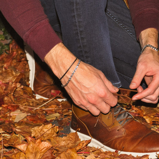 Person tying a brown leather shoe on a leaf-covered ground with a  black box chain 