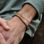 Close-up of a person's wrist wearing two bracelets one is a black rope and one is a gold box chain with a blurred background