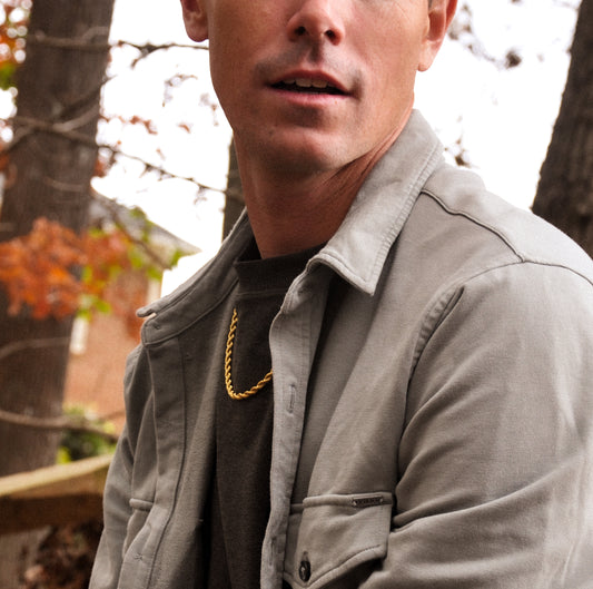 Man wearing a gold rope chain necklace, gray jacket with a blurred outdoor background