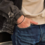 Person wearing a black jacket, black rope chain bracelet beige shirt, and blue jeans with a blurred background