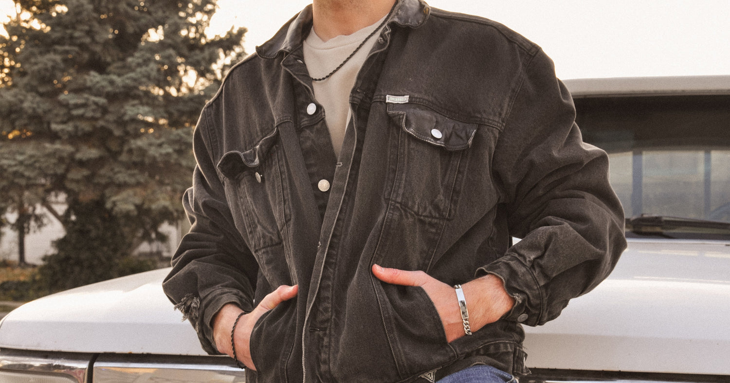 Person wearing a dark jacket and jeans with a black rope chain necklace and bracelet and silver snowflake obsidian bracelet standing in front of a white truck.