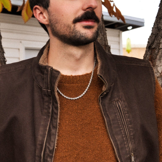 Man wearing a brown sweater and vest with silver rope chain necklace standing outdoors near trees.