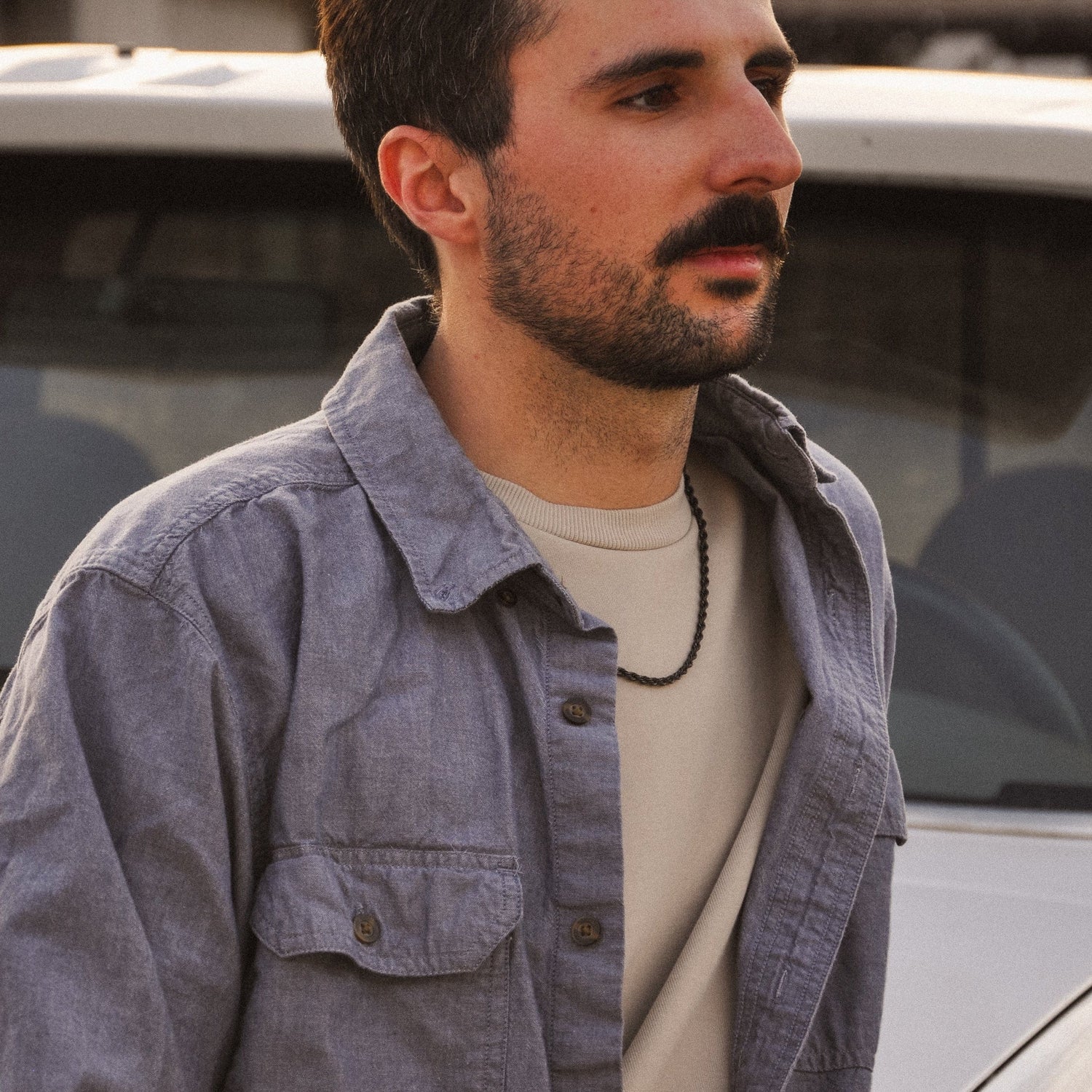 Man standing in front of a white van wearing a gray jacket with black rope chain necklace 